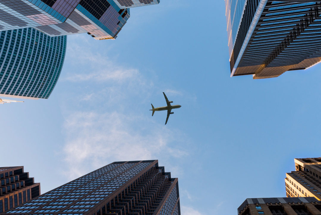 Blick in den Himmel auf ein Flugzeug, dass über mehrere Hochhäuser hinweg fliegt.