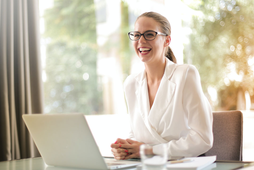 Fröhlich lächelnde Frau in weißem Kittel, die hinter einem Laptop an einem Tisch sitzt.